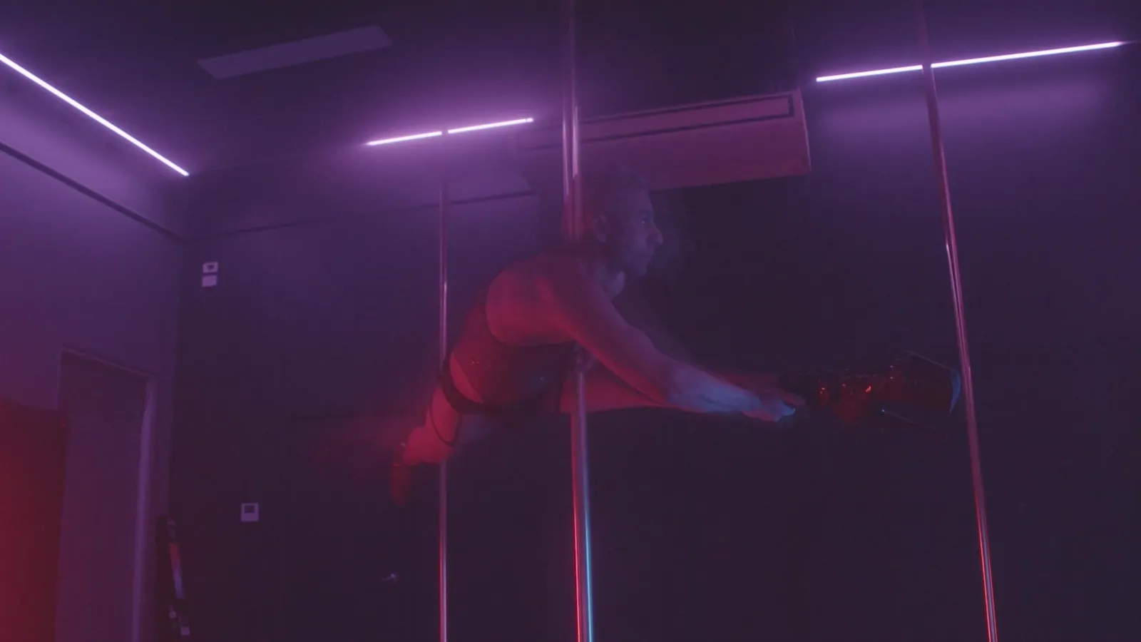Una persona de rojo hace una bandera horizontal en el pole en un estudio iluminado con neón