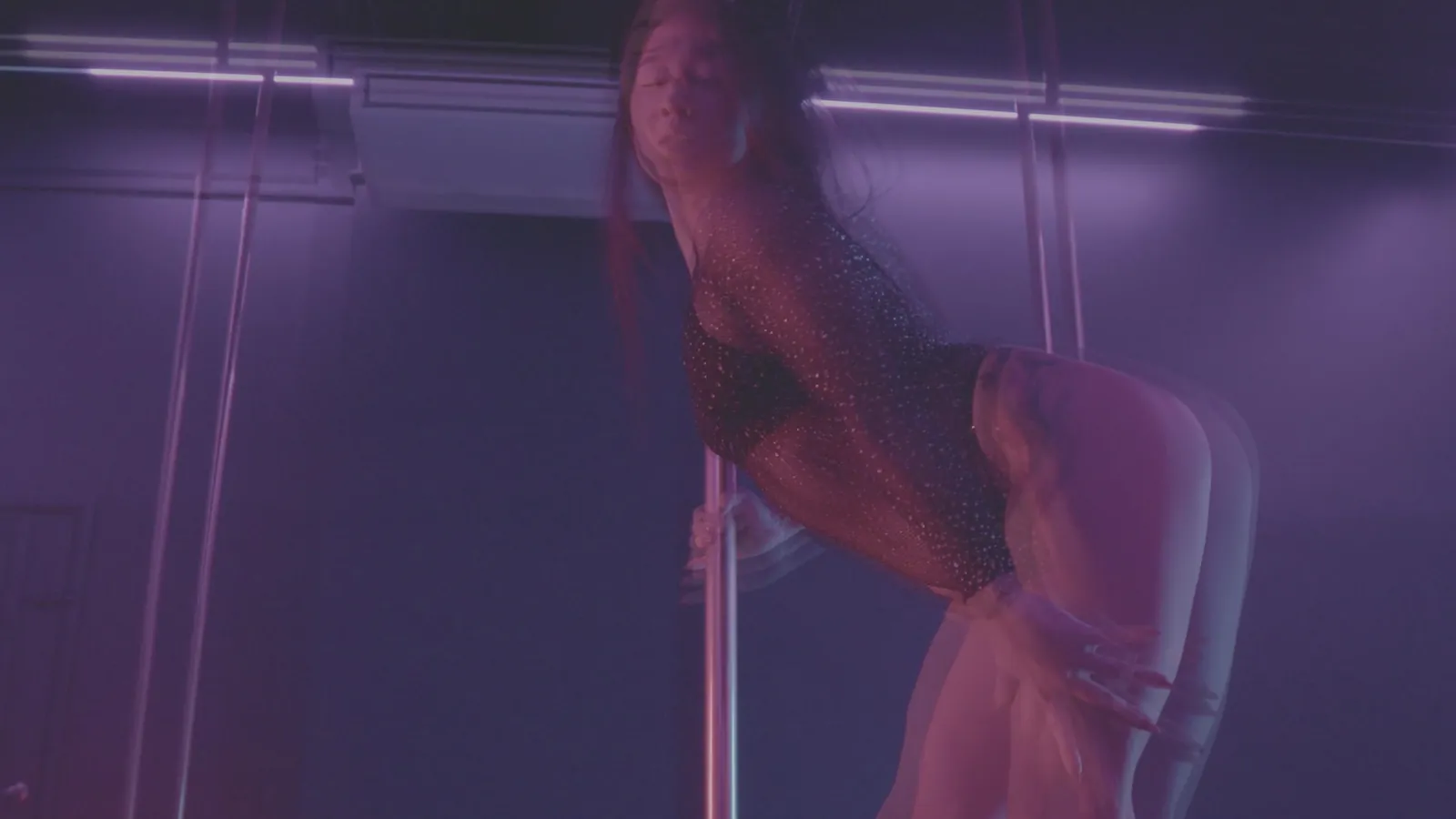 Una persona que baila pole en un estudio con luz morada, inclinada hacia atrás desde el pole
