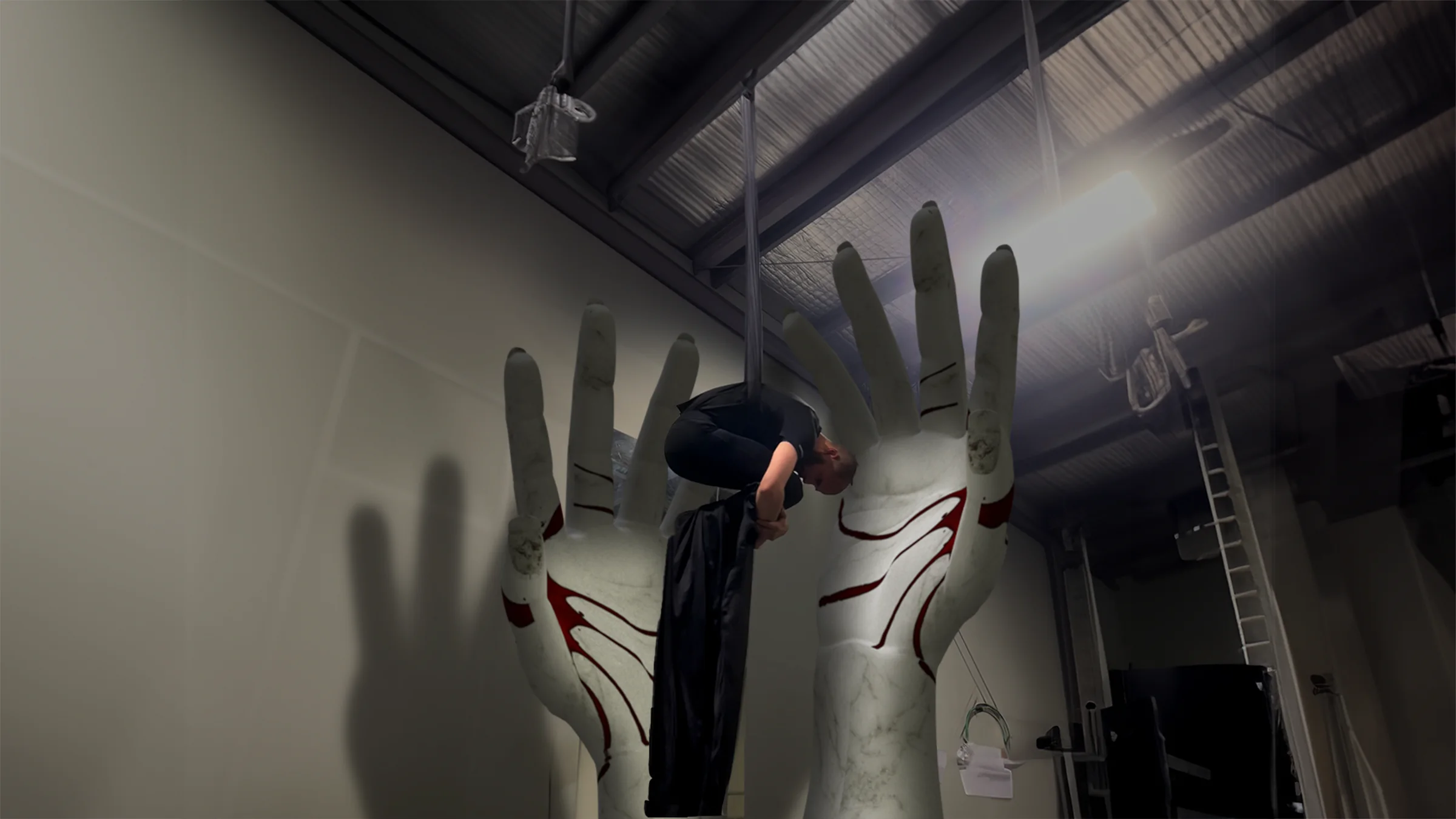 Composição de duas mãos gigantes de mármore com veios vermelhos saindo do chão de um estúdio, com uma pessoa artista aérea pendurada de cabeça para baixo entre elas