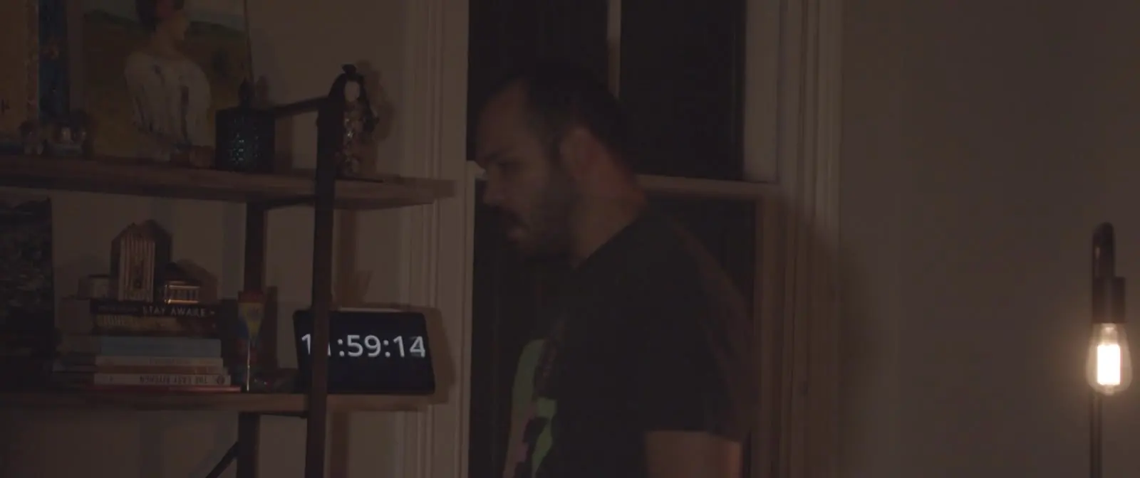 Um homem num quarto escuro iluminado por uma única lâmpada, ao lado de um relógio digital