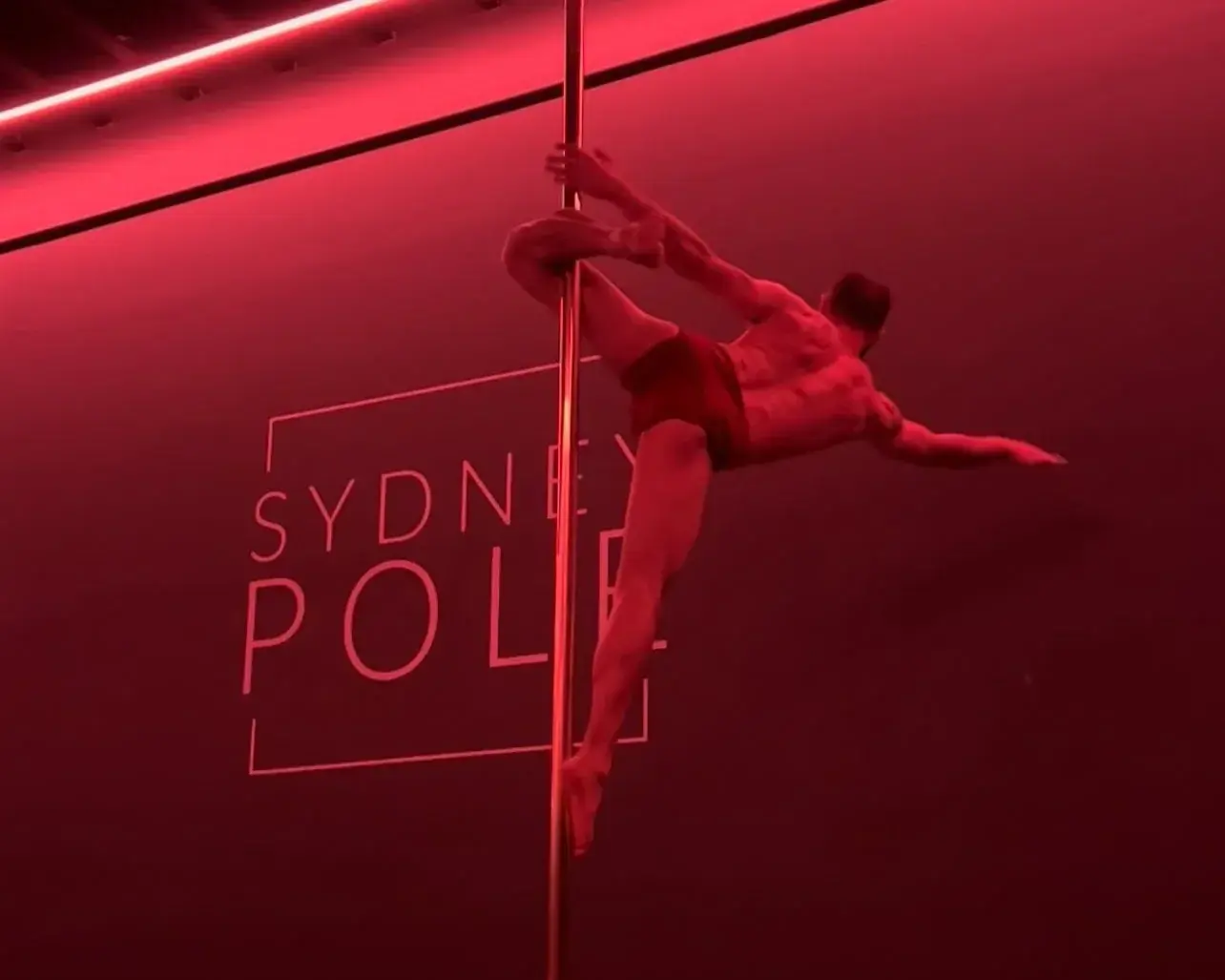 Uma pessoa fazendo parada de mão no pole em frente ao letreiro da Sydney Pole, iluminado de vermelho