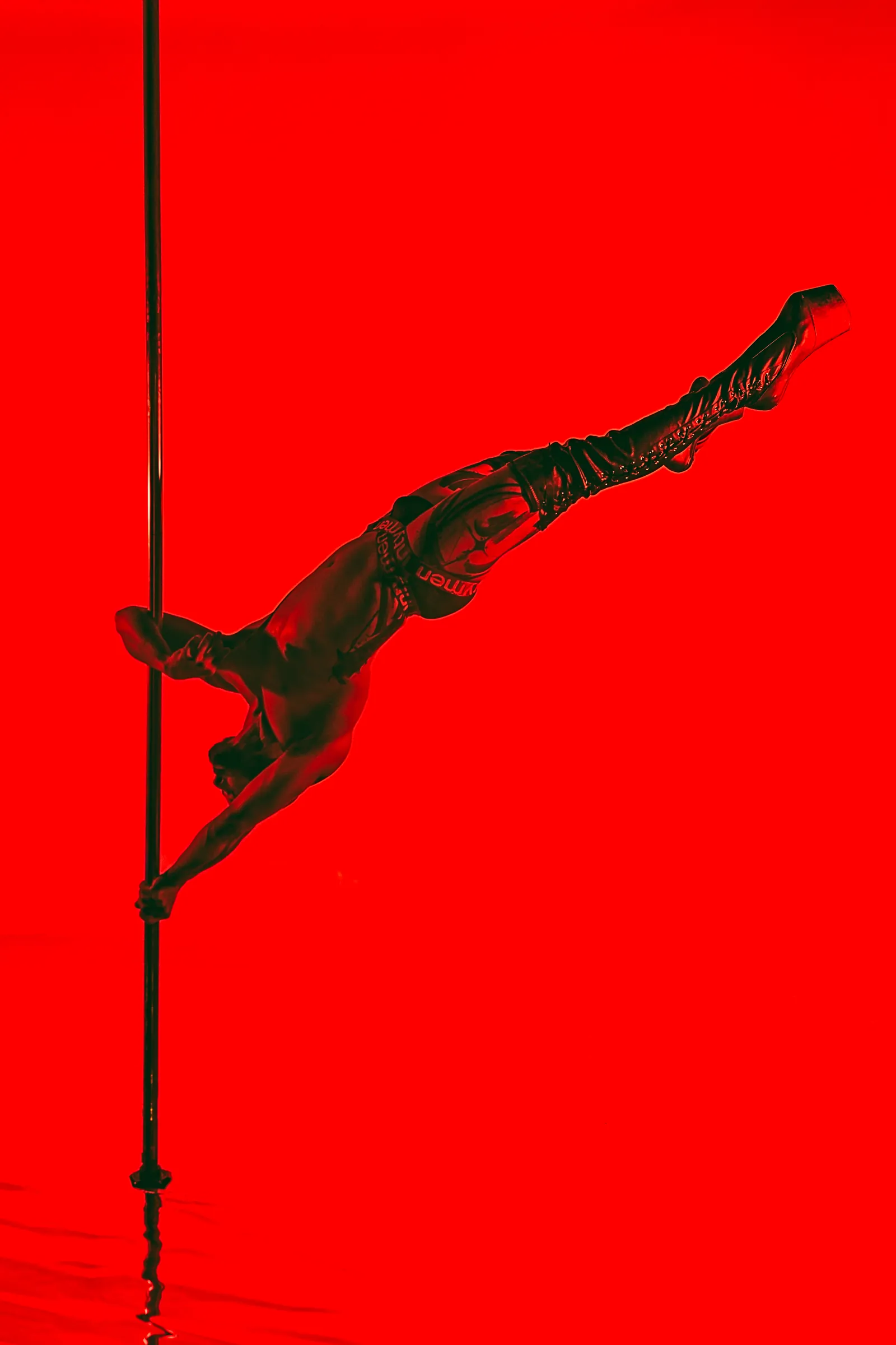 Augusto esticado de lado no pole, diante de um fundo vermelho profundo